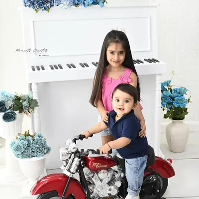 A fun and musical sibling portrait. The baby boy takes a ride on a toy motorcycle while his big sister stands by the piano, creating a playful and charming scene in our all-white studio setup.