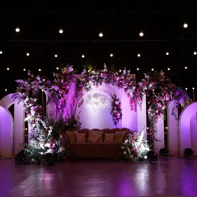 A dramatic and romantic reception stage featuring a lush floral arch in shades of purple and white. The custom neon sign and strategic uplighting create a vibrant, modern, and photo-ready backdrop.