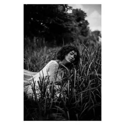 A dramatic black and white portrait in a field of tall grass. The high contrast and the model's pose create a moody, cinematic, and slightly mysterious feel.
