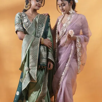 A beautifully composed shot of two models, showcasing a sage green silk saree and a lavender organza saree. This image is perfect for a lookbook cover, demonstrating how different pieces from a collection can be styled together.