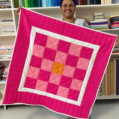 A happy student from our Beginner's Quilting Class holds up her first project, a vibrant pink and orange checkerboard quilt.