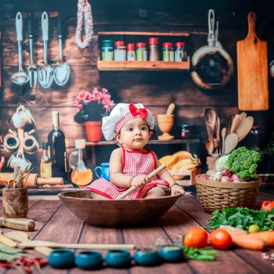 A curious little chef inspects her ingredients. The 'Little Chef' theme is perfect for sitter sessions, allowing babies to safely interact with various props like wooden spoons and fresh vegetables in a charming kitchen setup.