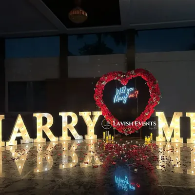 The perfect proposal setup. Giant illuminated "MARRY ME" letters, a heart-shaped arch made of red roses, and a neon "Will you Marry me?" sign create a breathtaking scene.