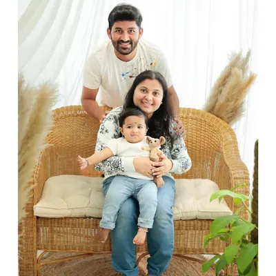 A warm and relaxed family portrait in a studio with natural-looking props. The parents and their baby are dressed in casual whites and blues, creating a cohesive and timeless look.
