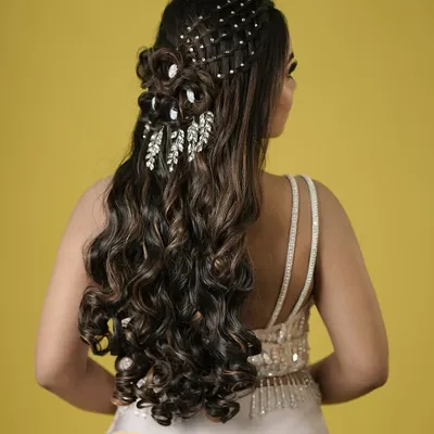 An intricate and glamorous half-updo with cascading curls, detailed with a pearl net and leaf-like crystal accessories. A true showstopper for a grand reception.