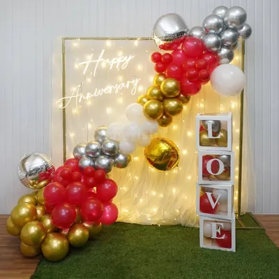 A popular choice for home anniversaries, this decor combines a trendy 'Happy Anniversary' neon sign with a backdrop of warm fairy lights. The diagonal balloon garland in red, gold, and silver adds a splash of color and fun.
