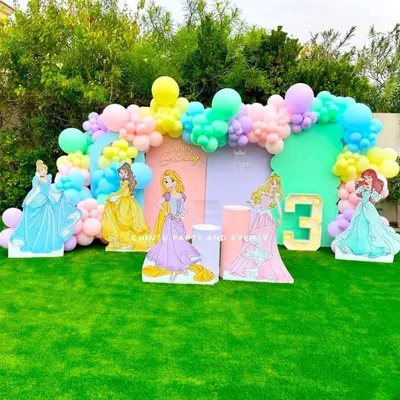A princess-themed birthday party featuring character cutouts of Cinderella, Belle, and others against a pastel backdrop. The setup includes a balloon arch and a light-up marquee number for the birthday girl.