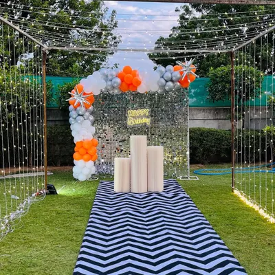 A stunning garden party entrance with a walkway leading to a silver shimmer wall, framed by fairy lights and an orange and silver balloon arch.