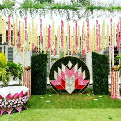 A modern and artistic naming ceremony setup in a garden. The centerpiece is a large circular frame with a geometric lotus pattern made of flower petals, flanked by green walls and hanging floral strings.