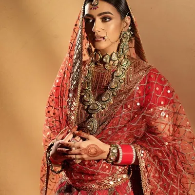 A traditional bridal portrait with a focus on rich, warm tones. The makeup complements the intricate red and gold outfit, featuring defined eyes and a matte-yet-radiant skin finish for a classic, royal look.