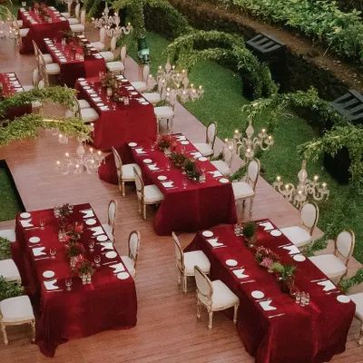 An aerial view of a spectacular engagement dinner setup in Goa. The rich red tables are arranged on a raised platform, surrounded by lush greenery and illuminated by elegant candelabras, creating a truly royal ambiance.