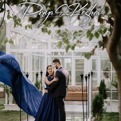 A dramatic pre-wedding portrait featuring a long, flowing blue gown. The movement of the fabric against the elegant white greenhouse creates a stunning, high-fashion look.