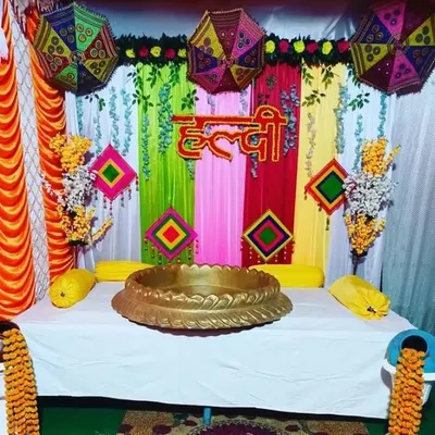 A vibrant and colorful backdrop for a Haldi ceremony. The setup features bright drapes, traditional umbrellas, marigold flowers, and a Hindi 'Haldi' sign at the center.