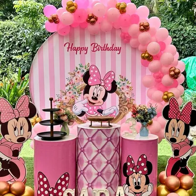 A classic Minnie Mouse birthday party setup in an outdoor garden setting. This decoration features a pink and white striped circular backdrop, a pink balloon arch with gold accents, and multiple Minnie cutouts.
