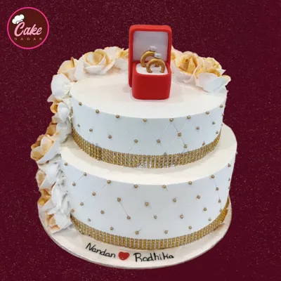 A beautiful two-tiered engagement cake in white and gold. The design features a quilted pattern, sparkling bands, and is topped with an edible ring box, symbolizing the special commitment.
