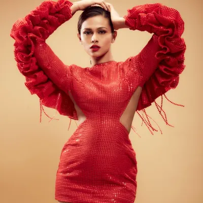 A full-length shot showcasing a dramatic red-on-red look for a high-fashion editorial. The makeup features a classic bold red lip and clean, defined eyes to complement the avant-garde styling of the structured gown.