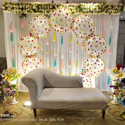 A unique Mehndi backdrop featuring an arch of decorative white umbrellas and colorful tassels. The floral border at the top and side arrangements complete this fun, mela-style setup.