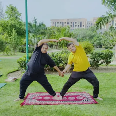 Two students form a heart shape with their arms while holding a partner standing pose in a park. This image perfectly captures the connection and playfulness we cultivate in our outdoor yoga sessions.