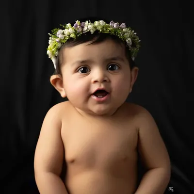 A simple, beautiful baby portrait against a classic black background. Wearing a delicate floral headband, her bright, curious eyes are the complete focus of the image.