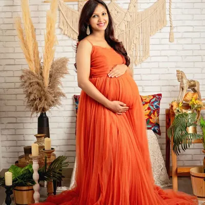 The vibrant orange gown against the rustic brick and macrame backdrop creates a stunning, warm portrait. Her smile says it all.
