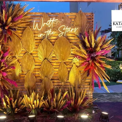 This romantic photo backdrop was designed for an engagement party. It features a wooden panel, a custom "Written in the Stars" neon sign, and dramatic floral bursts made with dried golden palm leaves and colorful accents.