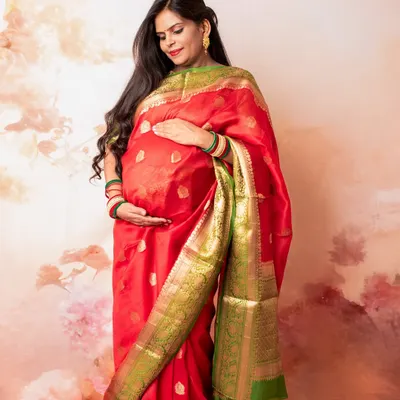 Embracing tradition and elegance, this studio maternity portrait showcases a stunning red and green silk saree. The soft, painted backdrop adds a touch of artistry to this classic look.