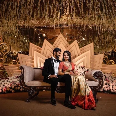 A formally dressed couple is elegantly posed on a luxurious chaise lounge against a grand, lotus-themed stage at their wedding reception. We specialize in capturing the scale and design of large-scale wedding events.