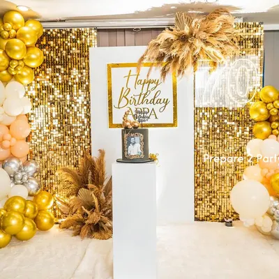 A chic and modern backdrop for a 70th birthday celebration. This setup features a gold shimmer wall, a white central panel with custom calligraphy, and organic balloon arches in gold, silver, peach, and white, accented with dried pampas grass.