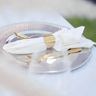 A close-up of a place setting featuring a hammered silver charger plate and gold-plated cutlery wrapped in a crisp white napkin with a gold ring.