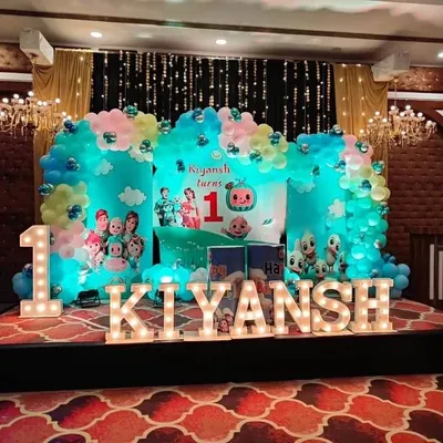 A complete Cocomelon stage setup for a first birthday party. This grand arrangement includes a large themed backdrop, marquee letters spelling out the name, a number '1' light, and a cloud-like balloon garland, perfect for a memorable celebration in a banquet hall.