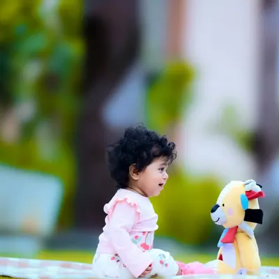 A baby sitting on a picnic blanket outdoors, having a sweet conversation with a teddy bear.