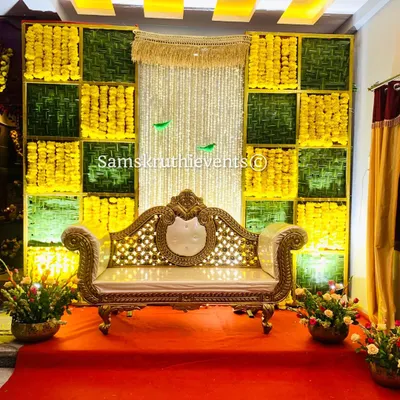 A classic Haldi or Mehendi backdrop featuring a checkerboard pattern of yellow marigolds and green woven leaves. The ornate gold and white sofa provides a perfect seat for the ceremony.