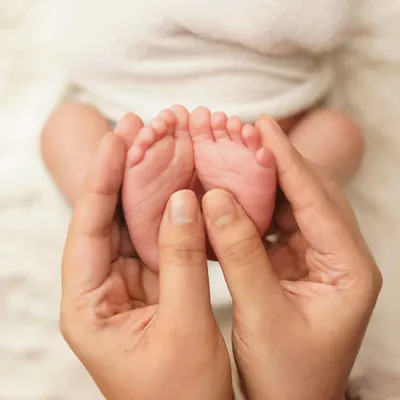 A close-up of a parent's hands gently holding their newborn's tiny feet. The image is shot from above against a soft white blanket, emphasizing how small and perfect they are.