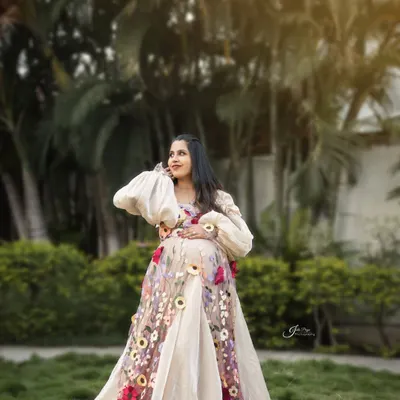 An outdoor maternity portrait glowing in the golden hour light. The expecting mother looks radiant in a beautiful floral embroidered gown, surrounded by lush greenery.