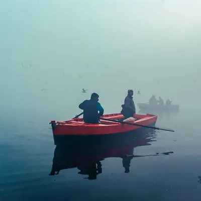 The bright red boat provides a striking pop of color against the foggy, muted tones of the Yamuna river. This contrast is what makes the scene so visually interesting.