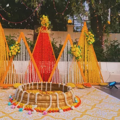 An outdoor Haldi ceremony setup with a backdrop of marigold strings in geometric patterns. The traditional brass urli in the center is filled with flowers for the ritual.