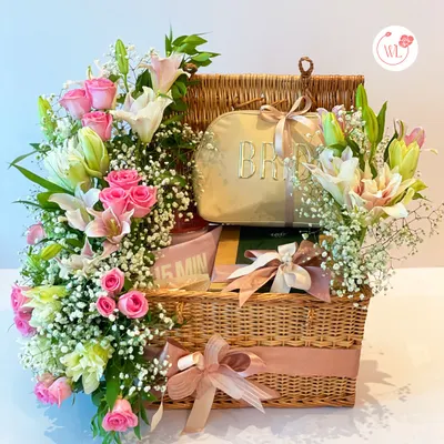 Elevate the bride's experience with this stunning essentials package. Housed in a classic wicker picnic basket, it's filled with curated gifts and adorned with an explosion of fresh pink roses and lilies.