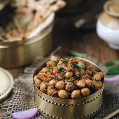 A moody, rustic shot of chole served in a traditional brass bowl. The dark lighting and textured surface bring out the rich colors of the curry, making it feel hearty and authentic.