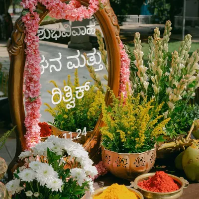 A personalized welcome mirror with the couple's names in Kannada script, surrounded by pink flowers and traditional elements like turmeric and kumkum.