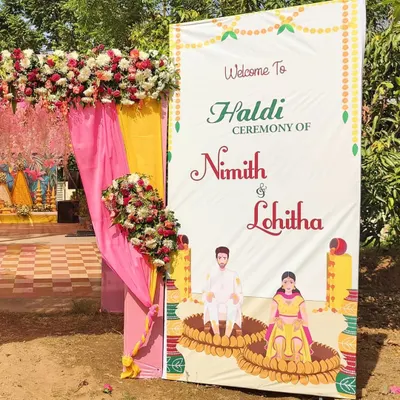 I design custom welcome boards like this one for a Haldi ceremony, featuring caricatures of the couple and event details. It's framed with matching floral arrangements.