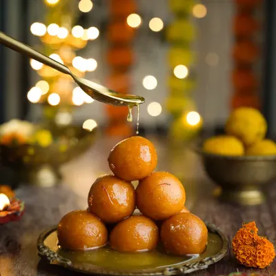 Happy Diwali. A classic festive shot of gulab jamuns stacked in a pyramid, with a spoon pouring syrup over them. The diyas and flower garlands in the background complete the scene.