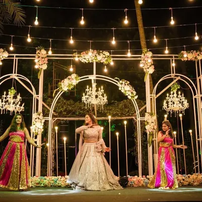 A spectacular stage for a Sangeet night performance. The design features white arches, multiple crystal chandeliers, and dramatic lighting to make every performance look like a professional show.