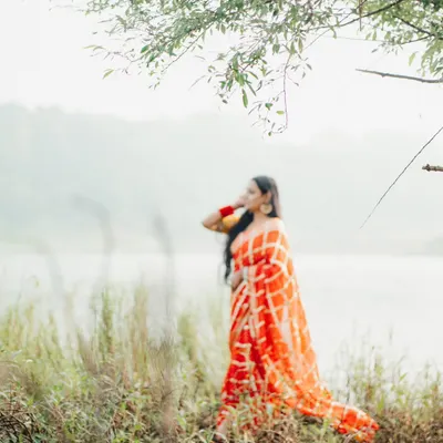 An artistic, soft-focus shot of a woman in a vibrant orange bandhani saree by a lake. This image has a dreamy, ethereal quality.