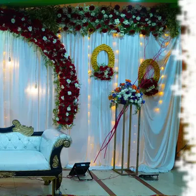 A classic and elegant stage design for a wedding or reception. This setup features a rich red and white floral arch, matching floral arrangements, and a backdrop of white curtains with fairy lights.