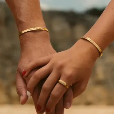 A couple holding hands, both wearing matching gold-plated cuff bracelets engraved with special coordinates. This is a beautiful way to commemorate a shared place or memory.