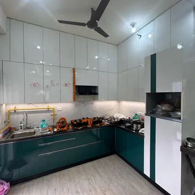 This recently completed 3BHK kitchen features a stunning combination of teal green and glossy white cabinets. The design includes a built-in chimney, under-cabinet lighting, and a marble-patterned backsplash for a clean, contemporary look.