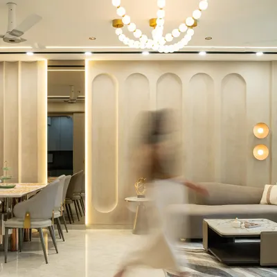 This luxurious living-dining space embraces fluidity through its curved wall paneling and layered lighting. A sculptural chandelier anchors the ceiling, while soft neutral tones, brushed gold accents, and plush textures create a seamless blend of modern sophistication and quiet opulence.
