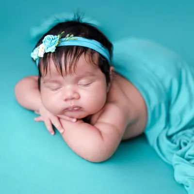 The 'chin on hands' pose is a newborn photography classic. We achieve this pose safely while the baby is in a deep sleep, using a vibrant teal backdrop and matching headband.