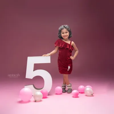 Celebrating five years with a stylish pre-birthday shoot. This young girl in a velvet dress poses confidently next to a large number '5' prop, surrounded by festive pink and white balloons.