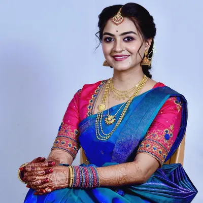 A studio portrait of a bride in a vibrant blue and pink saree. The makeup is classic and elegant, with a focus on defined eyes.